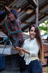 A beautiful young brunette girl looks after her horse in the stable. Feeding the stallion with some love.