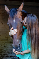 A beautiful young brunette girl looks after her horse in the stable. Kissing the stallion with love.