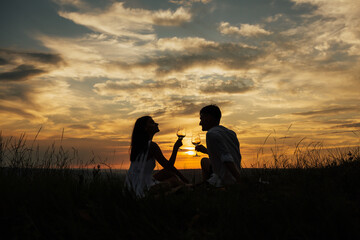 Obraz premium Silhouettes of young man and woman on holidays or honeymoon at amazing sunset sky. Man and woman clanging wine glasses with wine at sunset. Couple in love drinking champagne and celebrating their love