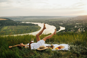 Gorgeous young girl in a white sundress lying on grass and having a picnic in a picturesque place....