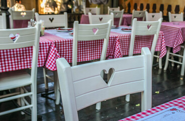 Empty white wooden chairs with cut out hearts and tables with red plaid tablecloths on a restaurant terrace