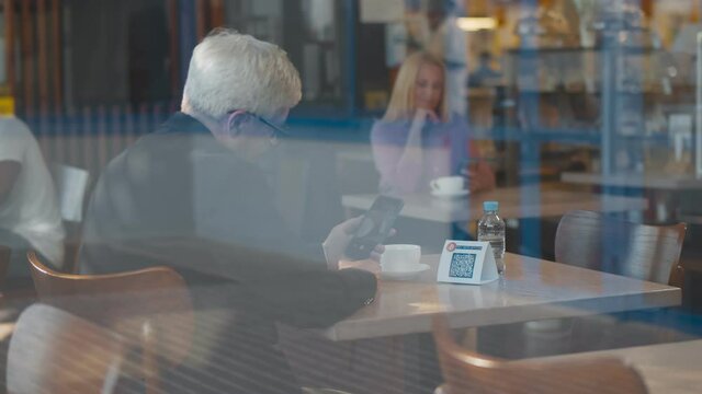 Side view through window senior man using smartphone app scanning qr-code to pay for order in cafe
