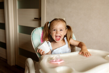 Little girl 2 years old with ponytail hairstyle plays with water, laughs and smiles, sitting in a highchair in the kitchen at home
