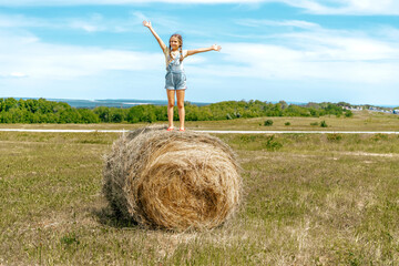 Naklejka na ściany i meble Cute little baby girl with pigtails and in a denim jumpsuit with open arms stands on a haystack in a field, the concept of summer vacation and freedom