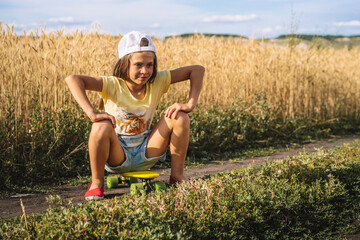 A little girl in a denim jumpsuit sits on a skateboard in the middle of a wheat field, a cheeky naughty teen © KseniaJoyg