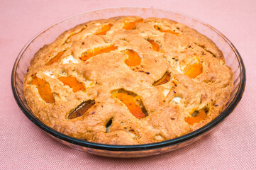 A large, round baked apple and apricot pie stands in a glass form on a table with a pink tablecloth. Delicious, fresh dessert.