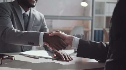 Midsection arc shot of African American and Caucasian male colleagues in formalwear shaking hands and discussing business while sitting at desk in office - Powered by Adobe