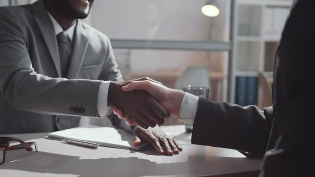Midsection arc shot of African American and Caucasian male colleagues in formalwear shaking hands and discussing business while sitting at desk in office