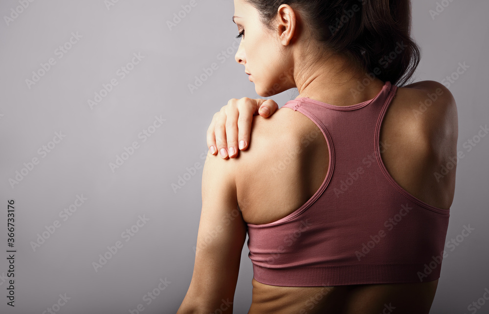 Wall mural Young strong sporty woman suffering from pain in shoulder in sport wear. Touching and massaging the hand. Sports exercising injury. Closeup portrait on purple background with empty copy space