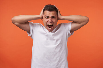 Irritated young brown-eyed short haired male covering his ears with raised palms and keeping mouth opened while screaming excitedly, isolated over orange background