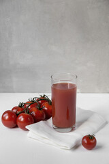 A glass with tomato juice stands on a white table next to ripe tomatoes lie on a branch from behind a light gray background
