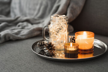 Living room decor: two burning aroma candles of orange color, pine cones and christmas lights in a glass jar. Hygge concept. Autumn - Winter season