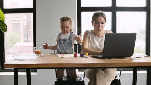 businesswoman typing on laptop while toddler child daughter is painting on paper iroi. freelancer mother working in home office. busy mom sitting with child at distant telework