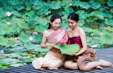 Beautiful Thai two women in thai traditional costume trying to roll up lotus flower while sitting on wooden path outdoors at lotus pond background. Asia young woman wearing Thailand 