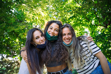 Portrait of a group of young millennials friends in a park at sunset with lowered face mask for the contagion from Coronavirus, Covid-19 - Beautiful women looking the camera and smiling