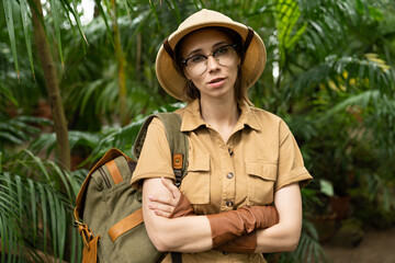 Fototapeta premium Close up portrait of woman botanist dressed in safari style in greenhouse. Naturalist in khaki clothes, leaver gloves with backpack looking at camera, arms crossed, surrounded by palms. 