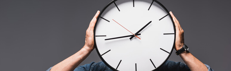 Panoramic crop of man holding clock near face isolated on grey, concept of time management