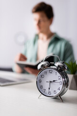 Selective focus of alarm clock on table and man holding notebook near laptop at home, concept of...