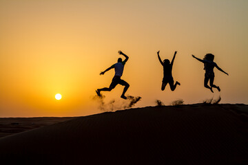 Fototapeta na wymiar Silhouette of happy traveling people jumping on sand dune and throwing sand in the air in golden sunset hour.