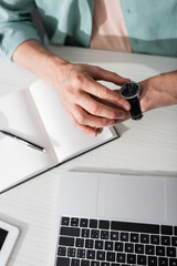 Cropped view of freelancer checking time on wristwatch near notebook and laptop on table, concept...