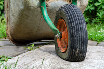 A wheel of an old and rusty garden wheelbarrow, deflated by a puncture, standing on a concrete...