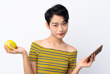 Young Asian girl having doubts while taking a chocolate tablet in one hand and an apple in the other