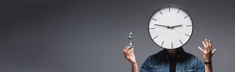 Panoramic shot of man with clock on head holding hourglass and gesturing on grey background, concept of time management