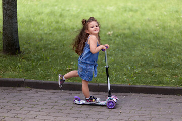 Naklejka premium Beautiful little girl rides a scooter in the park. Beautiful brunette girl.