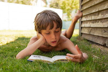 boy enthusiastically reading a book lying on the grass in his garden, vacation, back to school,...