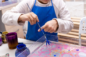 Fototapeta na wymiar Ebru - Traditional art of origin from Turkey. The process of transferring an image to a sheet of paper. A 6-year-old girl is painting in the workshop. Close up hands with ebru tool.
