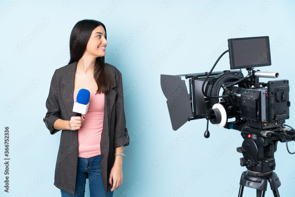 Wall mural Reporter woman holding a microphone and reporting news isolated on blue background looking side