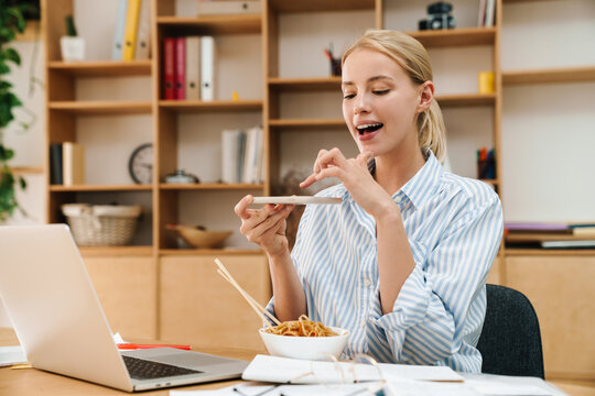 Image of woman taking photo while eating noodles and working with laptop