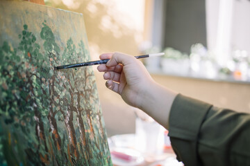 Woman's hand holding paintbrush and painting. Human's hand painting picture on canvas on tripod in yard with blurred house in background.