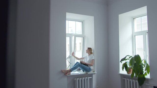 Girl sitting on windowsill in room with white walls with cat on her lap taking a picture or shoots video on her phone for social networks. Camera in motion