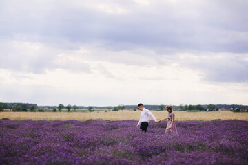 Young couple in love hugging and walking in a lavender field on summer clody day. girl in a luxurious purple dress and with hairstyle