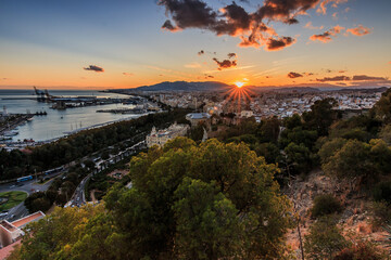 Sunset over the rooftops of the city of Malaga. Spanish coast in Andalusia with sun rays and clouds. City view on the Costa del Sol from an elevated position with harbor, streets and mountains