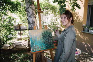 Beautiful female artist holding paint brush in front of painting. Young woman holding paint brush and looking at camera while standing in outdoor atelier.