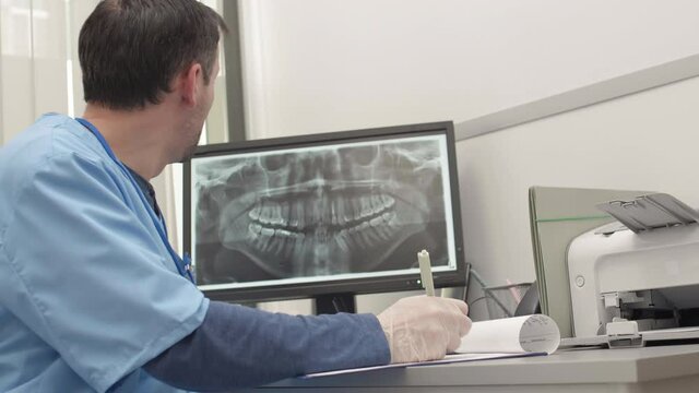 Medium shot of middle-aged male dentist wearing medical overall sitting at desk in the office, examining x-ray image of jaw on screen and filling in patient card