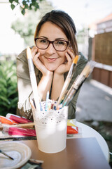 Cute woman with glasses posing and looking at camera. Girl posing while leaning against table with paint brushes in pot and paint tubes outdoor.
