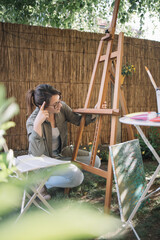 Woman squatting in garden while looking at wooden stand. Portrait of girl setting canvas stand for...