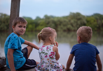 three children sit with their backs on the pier and look at the river. two brother and sister. blue sky. copy space, blurred background.