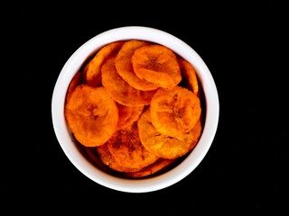 Orange color banana chips with hot red chili flavor in a white bowl against black background with copy space