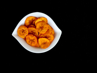 Orange color banana chips with hot red chili flavor in a white bowl against black background with copy space