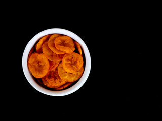Orange color banana chips with hot red chili flavor in a white bowl against black background with copy space