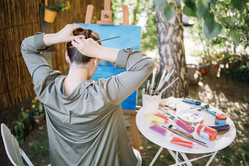 Brunette woman making hair bun using paintbrush. Back of woman making hairstyle with brush while sitting in outdoor art studio.