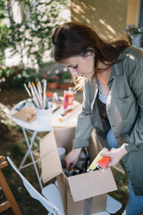 Young woman picking paint tubes from box. Side view of woman holding paint tubes and bending over box in backyard.