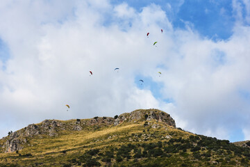 Fototapeta na wymiar Many paragliders on bright colored parachutes fly together over the mountain - Group of paragliders flying in sunny day against the background of clouds - Adventure, sport, and freedom concept