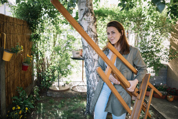 Attractive woman carrying canvas stand in backyard. Female painter holding tripod while walking in yard with flower, plants and tree.