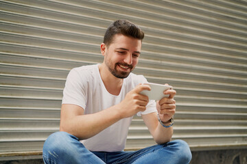 The young man sitting in front of the gray background is playing games with his mobile phone, holding his phone horizontally. The man is smiling while playing games with his mobile phone.   