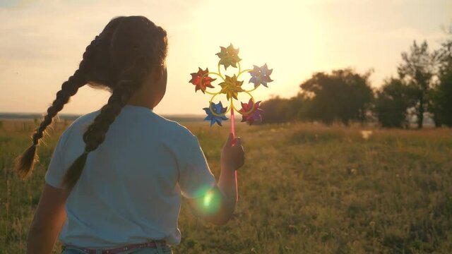 A happy, sweet girl runs across a field at sunset with a windmill in her hand. A child plays in the open air on a meadow.